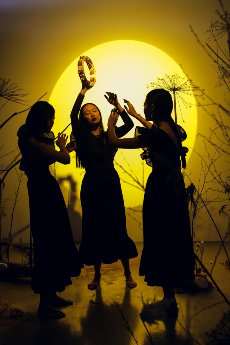 Three women dance with a tambourine under dramatic studio lighting, creating striking silhouettes.