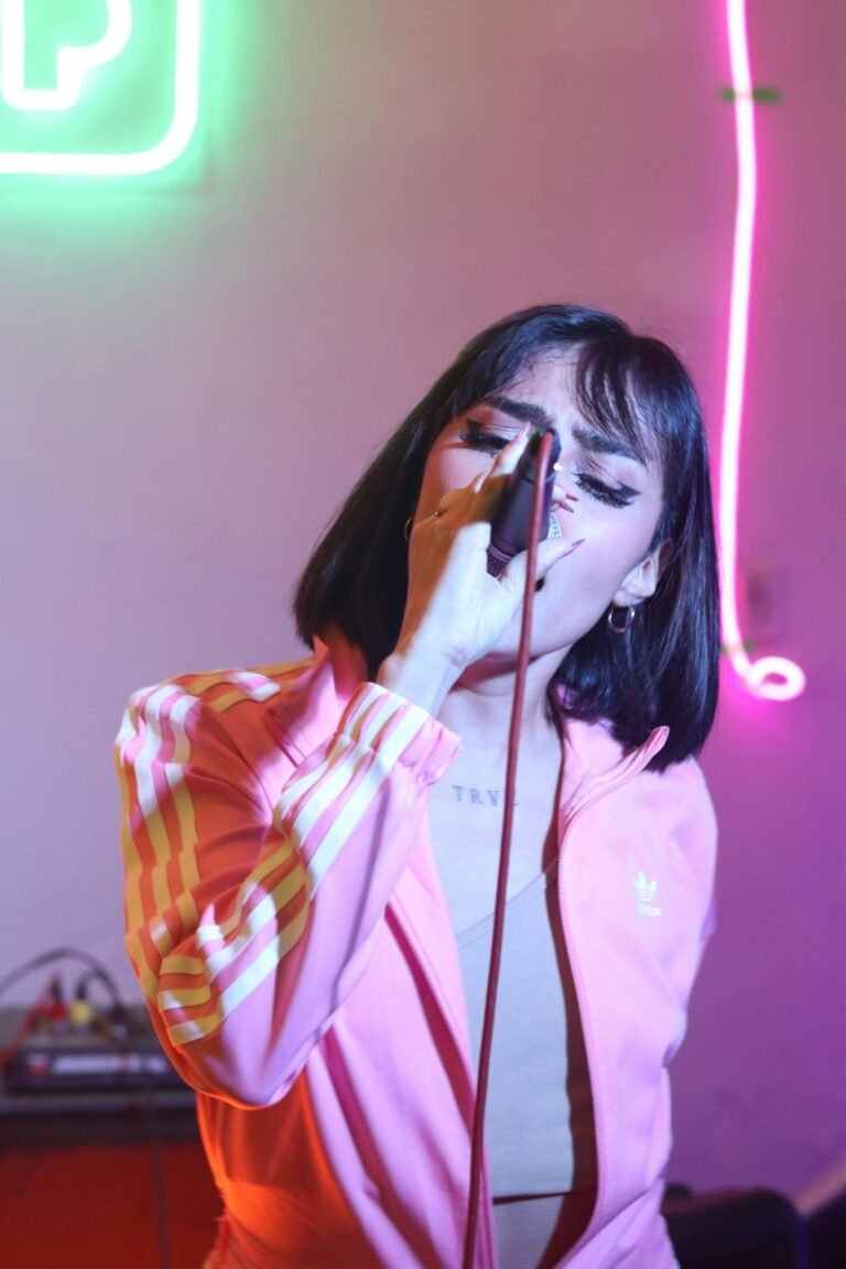 Woman in pink jacket singing into a microphone with neon lights behind her.